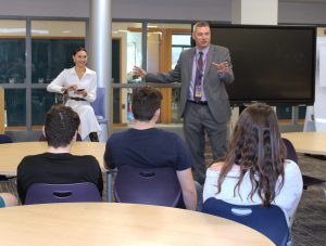 a woman in a white dress sits in a high top chair and speaks to a room full of students