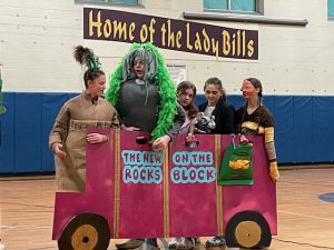 students perform a skit in costume and stand behind a school bus that they built