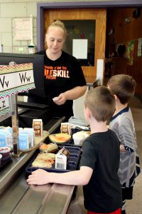 a woman stands at a cash register and young children go through a cafeteria line