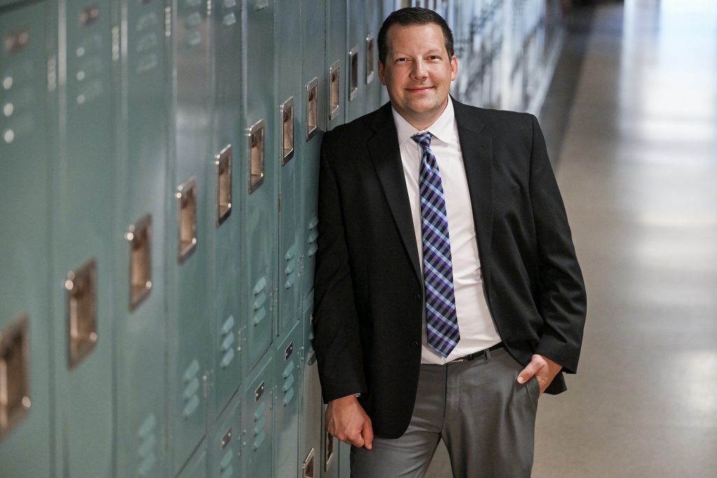 a man wearing a dark coat, white shirt and tie leans against at row of lockers in a school hallway