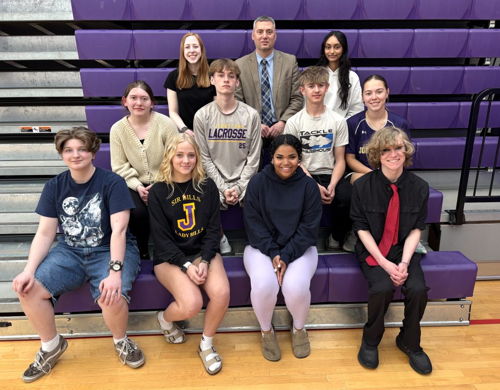 ten young adults sit on purple gym bleachers with an adult man
