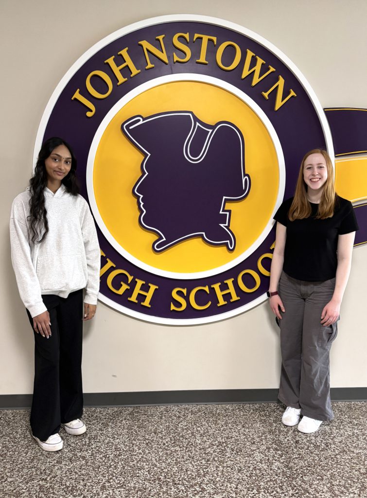 two young women stand in a hallway in front of a purple and gold school logo