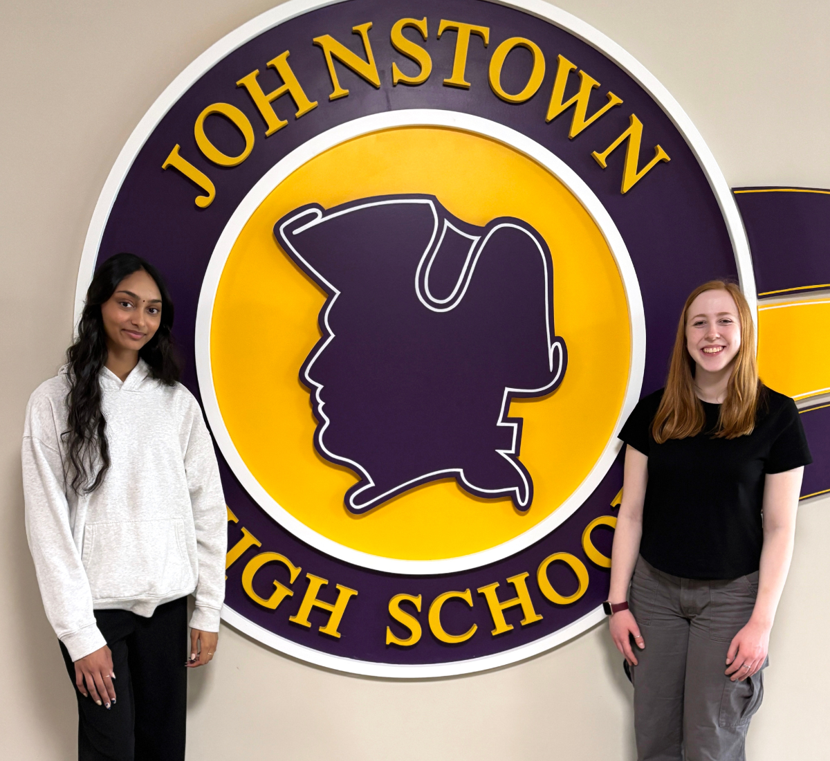 two young women stand in front of their purple and gold school logo
