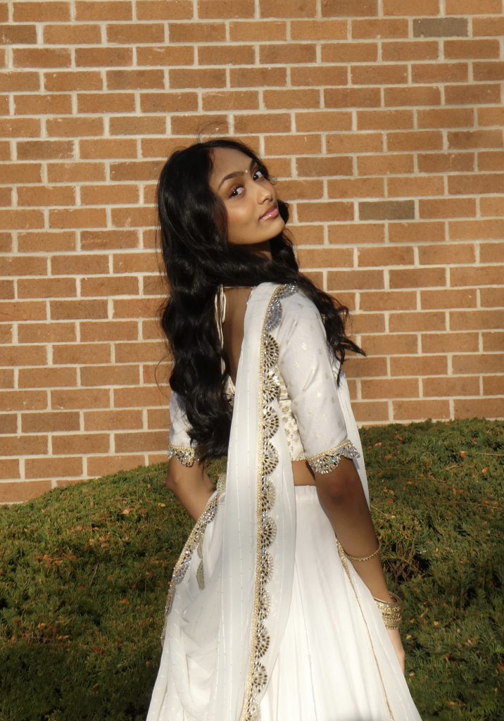 a young woman wearing a decorative white dress poses outside in front of a brick wall