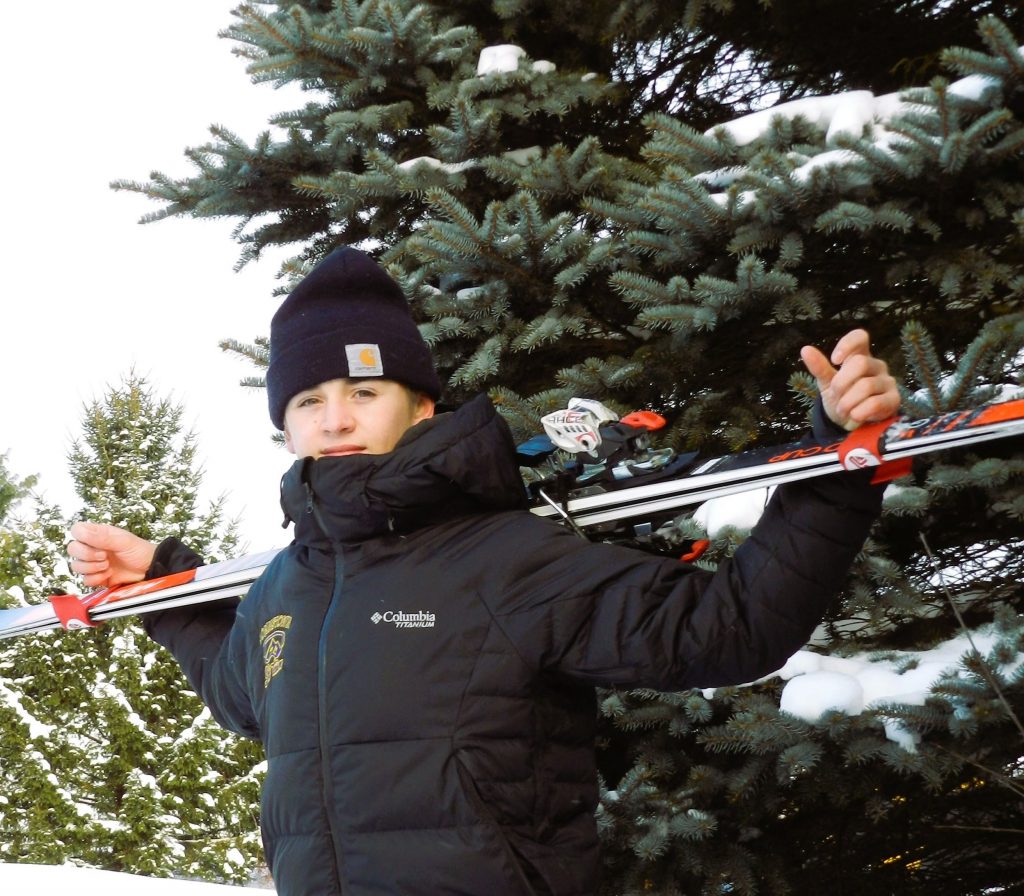 a young man wears a black winter jacket and hat while holding skis in front of a pine tree