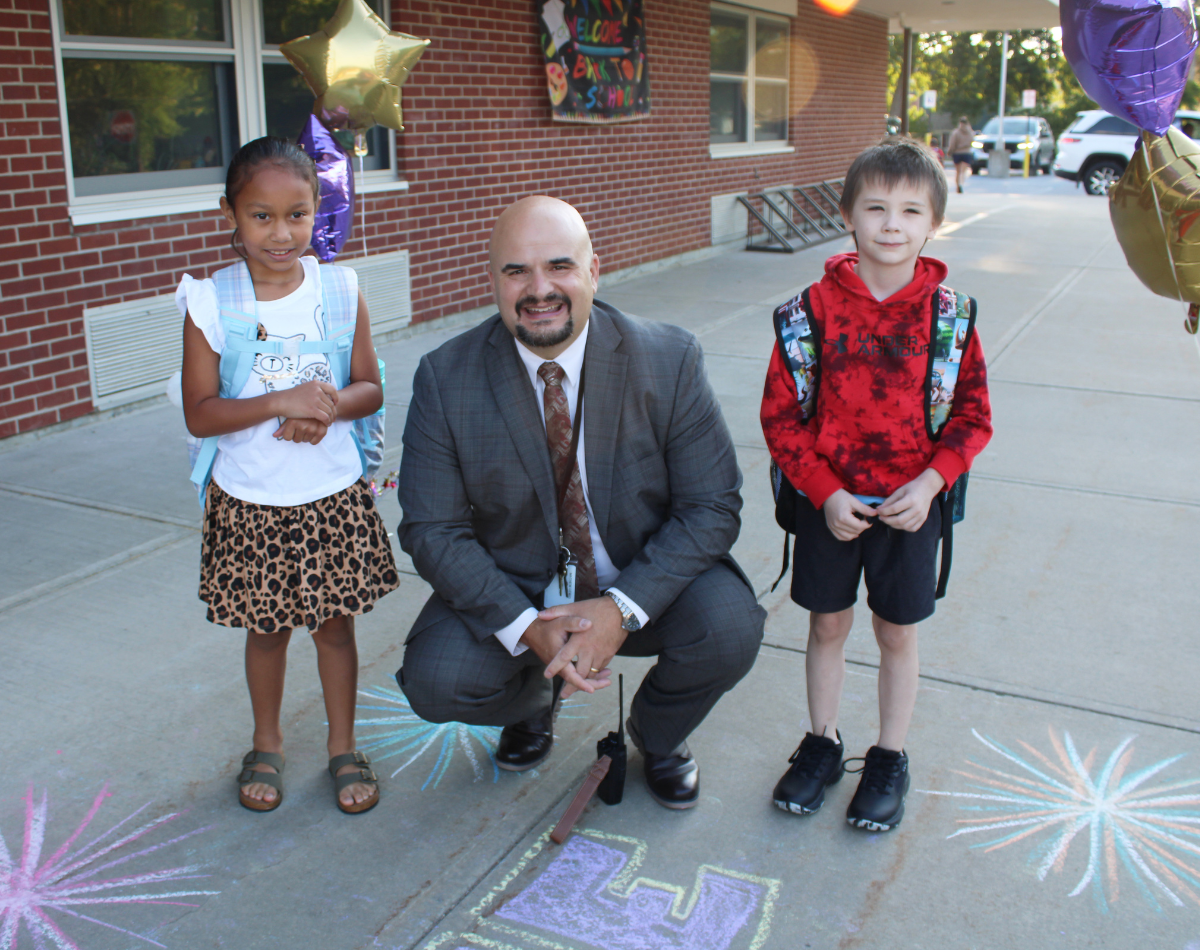 a bald man stands in between two young students outside on the sidewalk