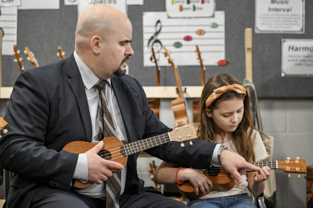 a bald man wearing a suit and tie teaches a young girl how to play the ukelele.