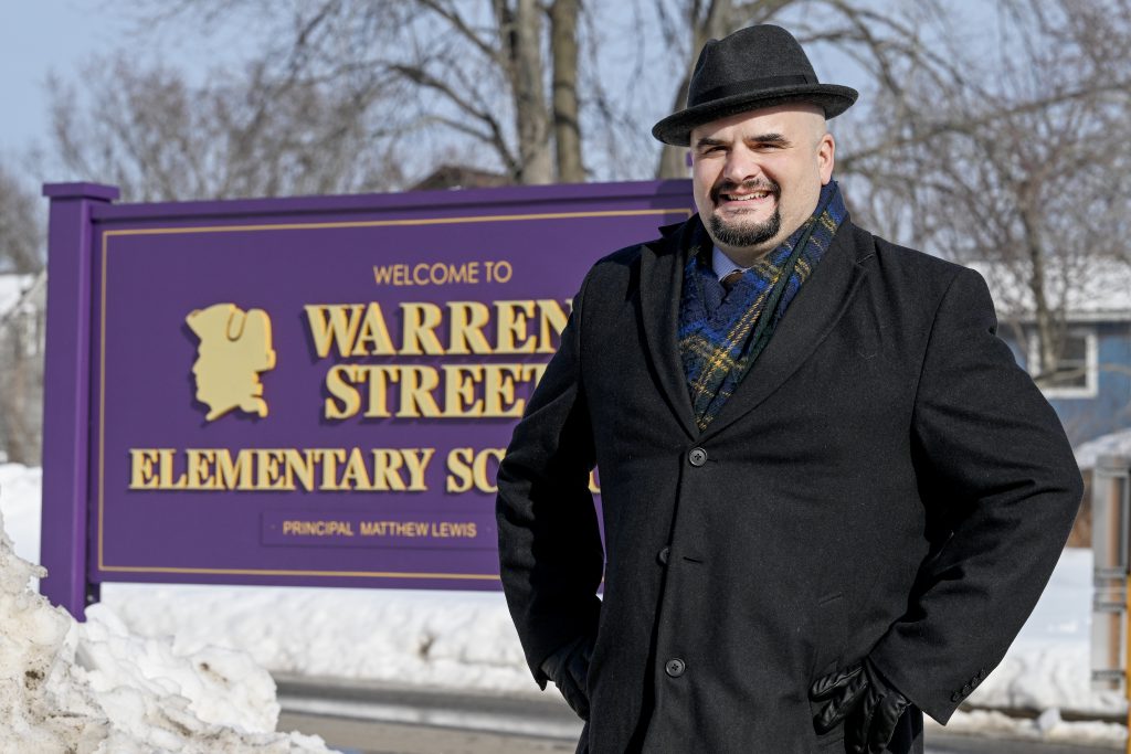 a man wearing a winter coat and hat stands outside in front of a purple school sign that reads "Welcome to Warren Street Elementary"
