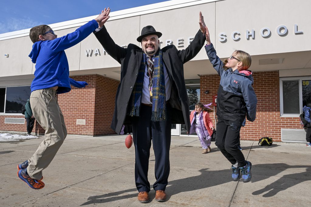 hat stands outside a school building and gives high fives to students as they jump in the air
