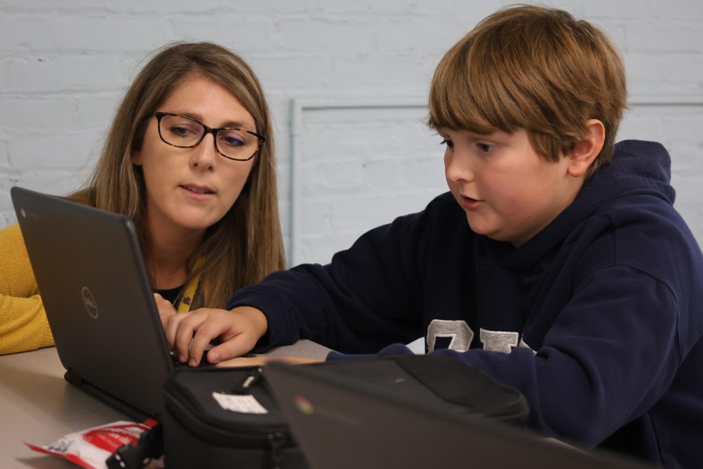 a woman with glasses assists a young student on their Chromebook