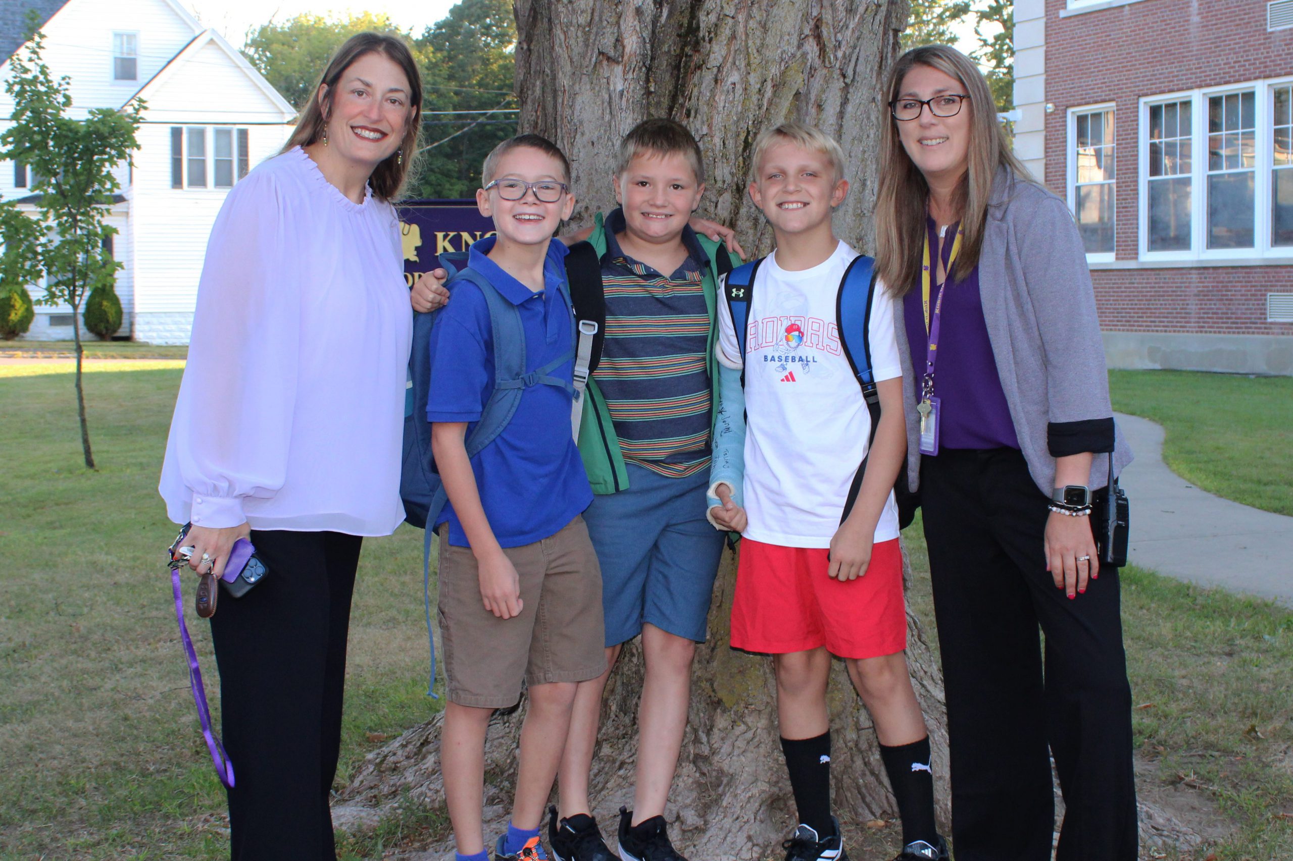 two adult women stand next to three young boys outside of a school, next to a tree