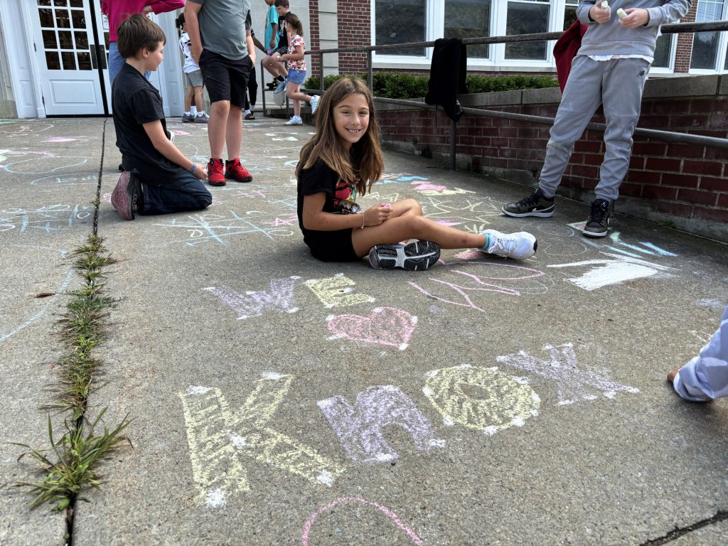 a young girls sits on a sidewalk outside of a school building and writes "We love Knox" with chalk