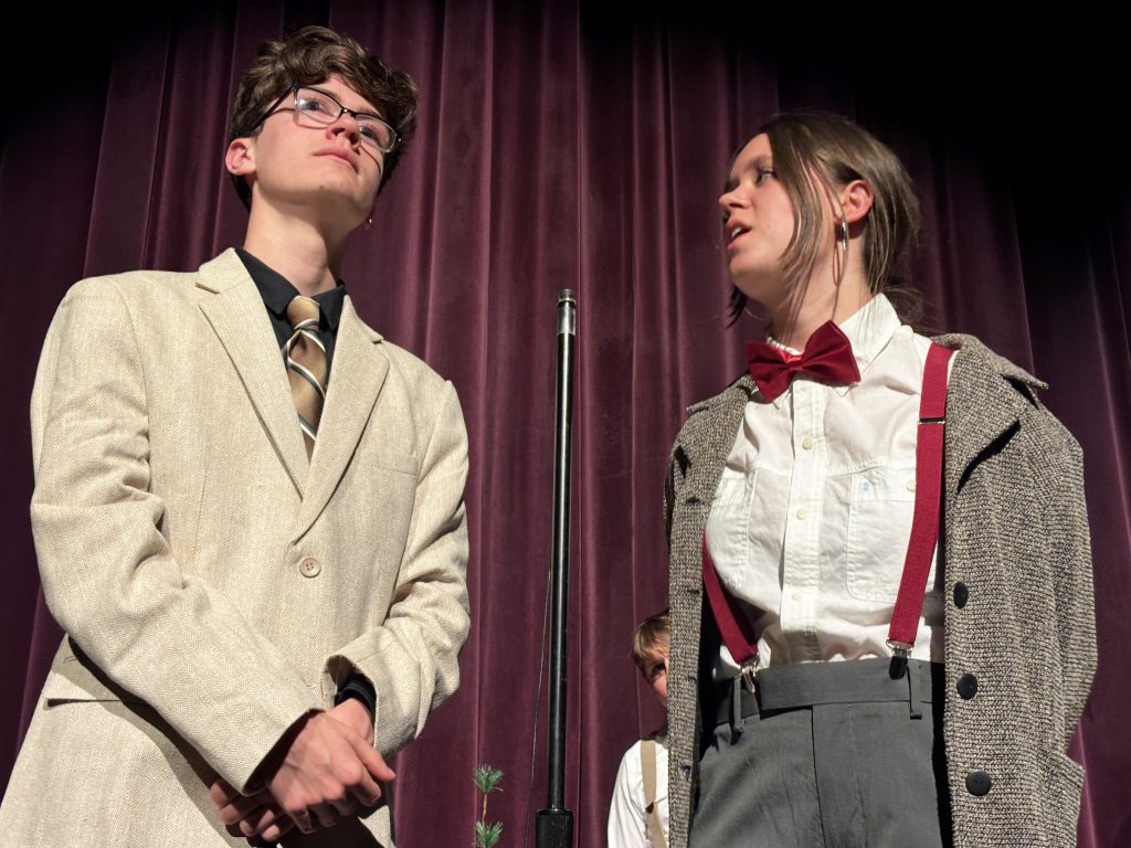a young man with a suit and tie stands next to a young lady wearing a bowtie on a stage