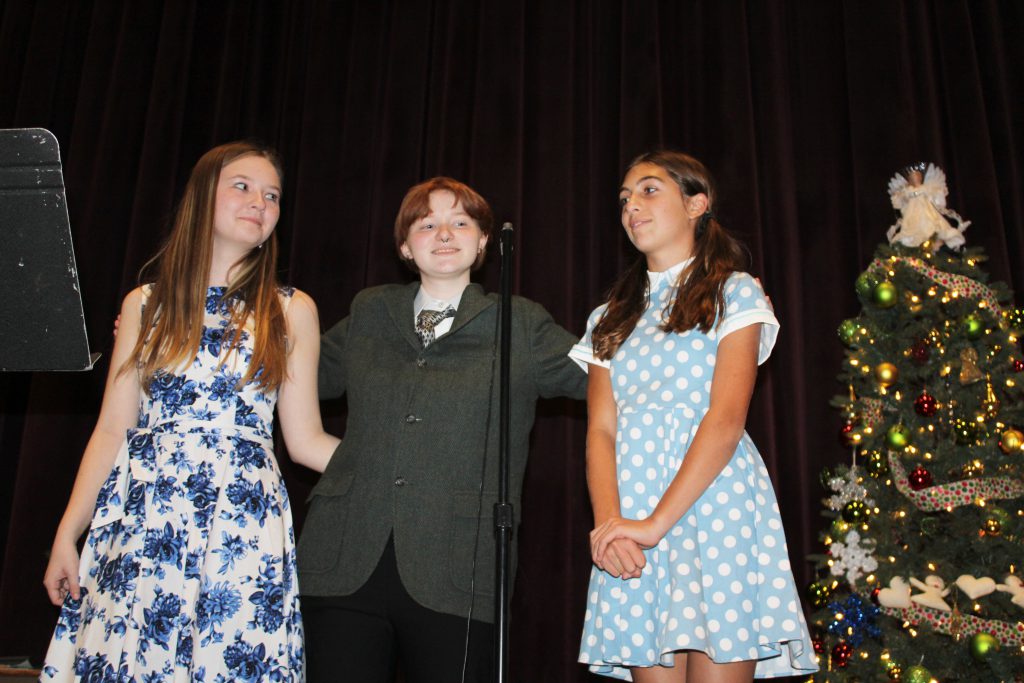 three young ladies stand on a stage and talk into a microphone in front of a Christmas tree