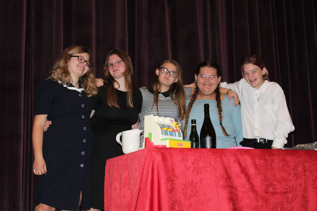 five young ladies stand on stage behind a red table