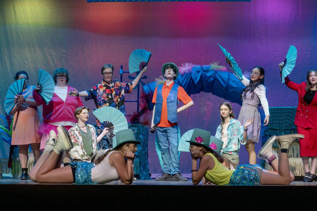 students in colorful costumes perform a scene from a musical on stage