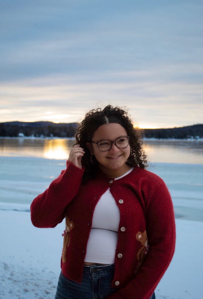 a young girl wears a red cardigan standing in front of a lake