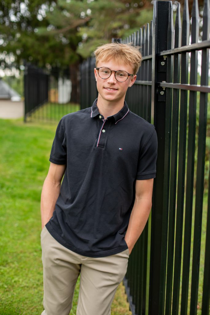 a young man with a black button down shirt and glasses leans against a black fence outdoors
