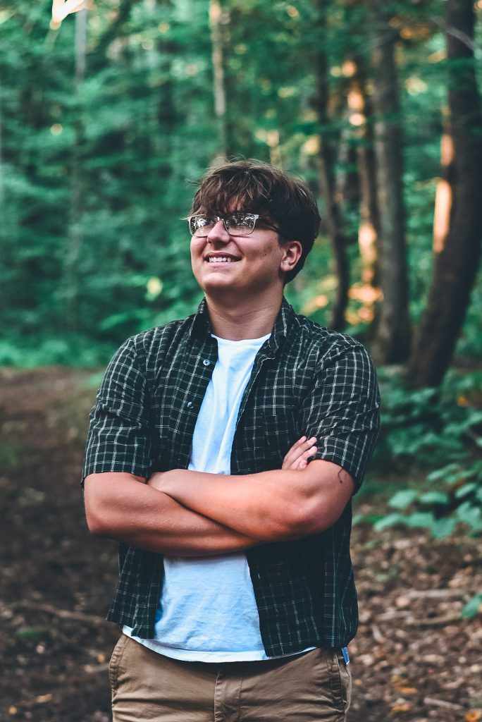 a young man with a plaid shirt poses outside among trees in nature