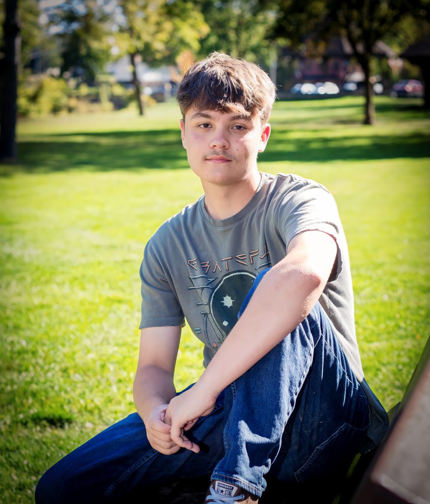 a young man wearing a grey t-shirt poses outside in the lawn