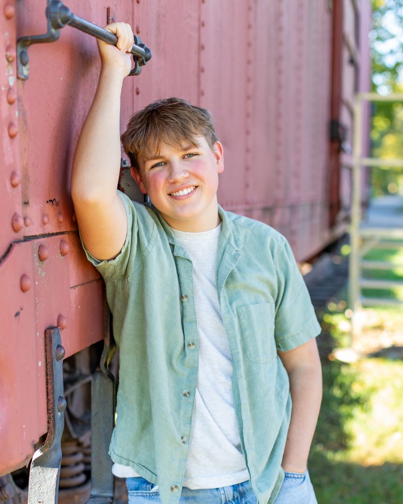 a boy wearing a light green button down shirt poses outside holding onto a train