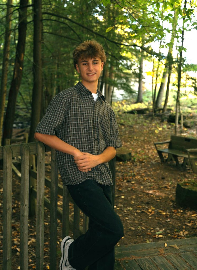 a young man wearing a plaid shirt leans against a wooden railing outside in nature