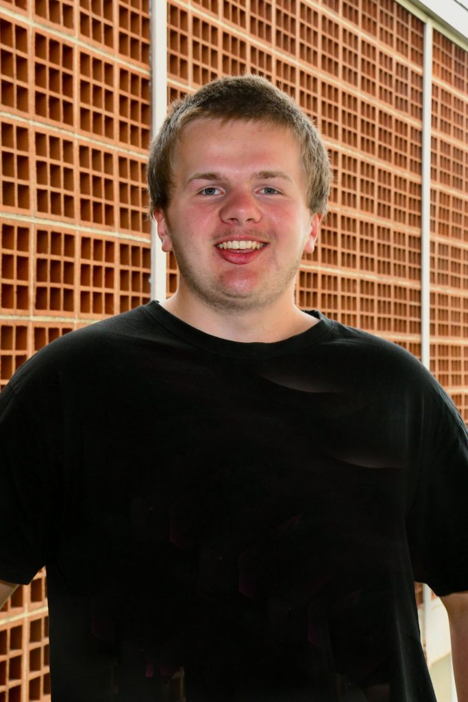 A young man wearing a black t-shirt stands in front of a geometric wall