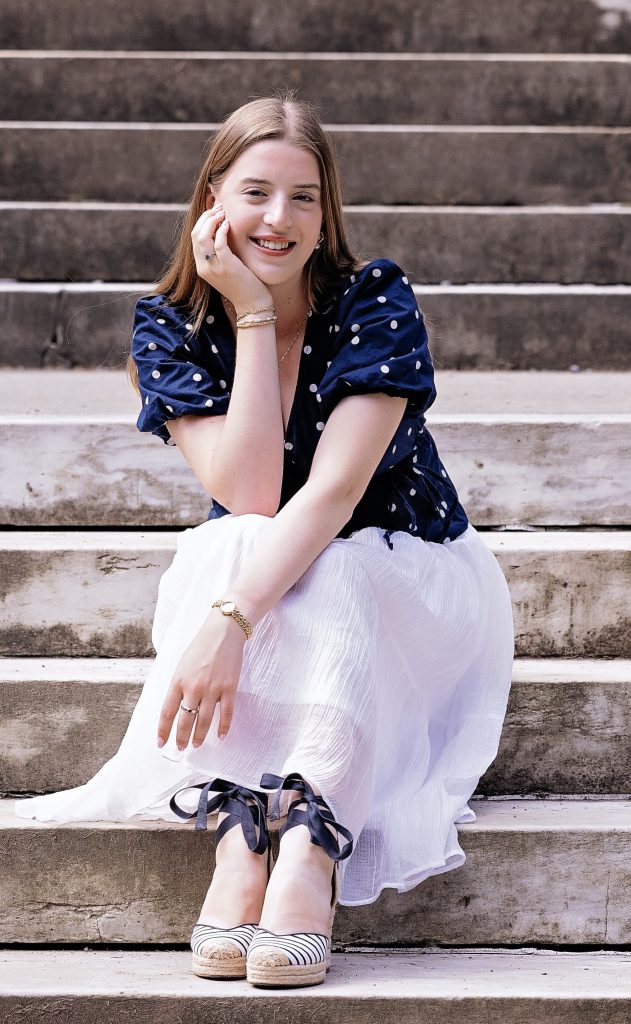 a young woman with a blue polka dotted shirt and white skirt sits on cement stairs outside